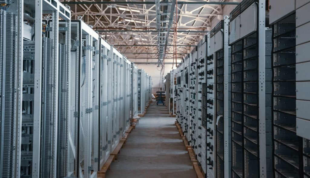 Rows of electrical switchgear and power distribution cabinets lining a long aisle inside an industrial data center building, with exposed steel roof trusses overhead, during equipment installation.