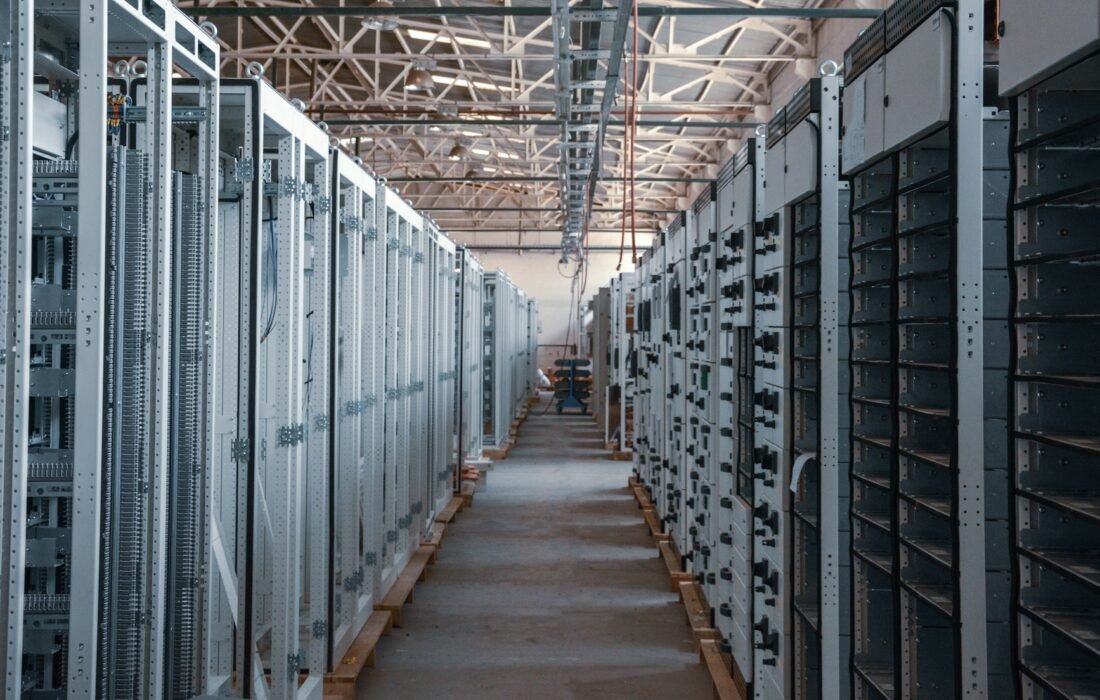 Rows of electrical switchgear and power distribution cabinets lining a long aisle inside an industrial data center building, with exposed steel roof trusses overhead, during equipment installation.