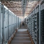 Rows of electrical switchgear and power distribution cabinets lining a long aisle inside an industrial data center building, with exposed steel roof trusses overhead, during equipment installation.