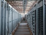 Rows of electrical switchgear and power distribution cabinets lining a long aisle inside an industrial data center building, with exposed steel roof trusses overhead, during equipment installation.