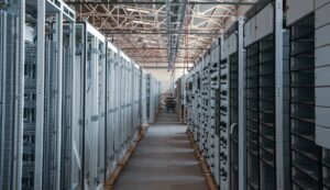 Rows of electrical switchgear and power distribution cabinets lining a long aisle inside an industrial data center building, with exposed steel roof trusses overhead, during equipment installation.