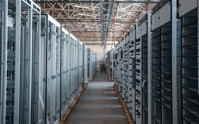 Rows of electrical switchgear and power distribution cabinets lining a long aisle inside an industrial data center building, with exposed steel roof trusses overhead, during equipment installation.